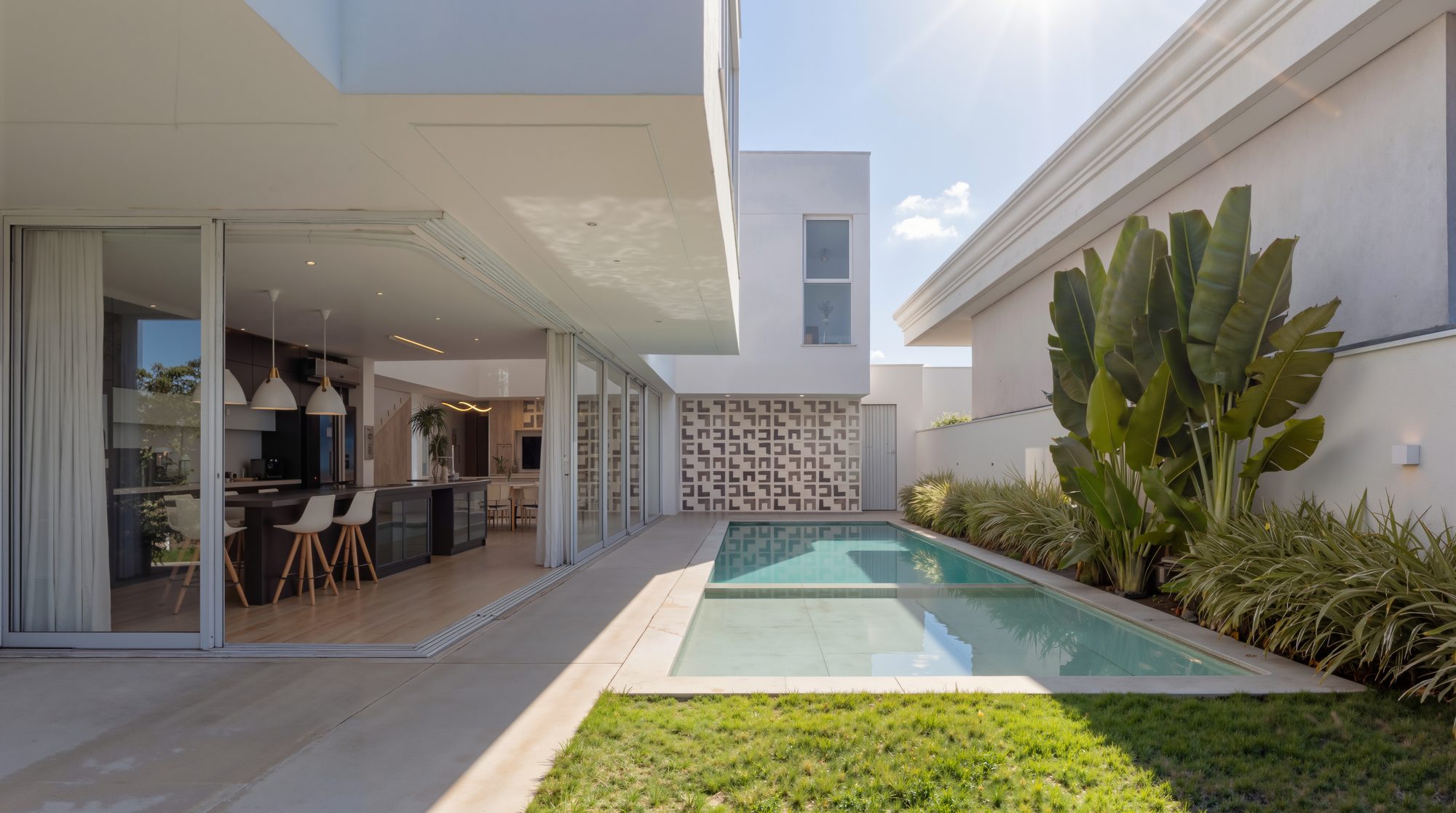 Área de lazer de residência contemporânea durante o dia: cozinha integrada aberta para a piscina, cobogós ao fundo e jardim tropical